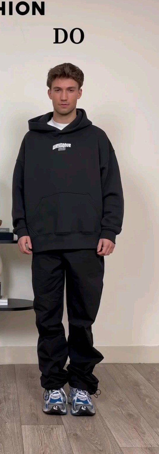 Man in the same black oversized hoodie with relaxed wide-leg trousers that drape over his sneakers