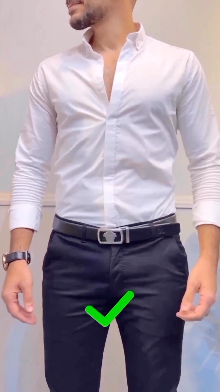 Man in an open-collar white button-up shirt tucked into dark tailored trousers with a black belt and green check overlay