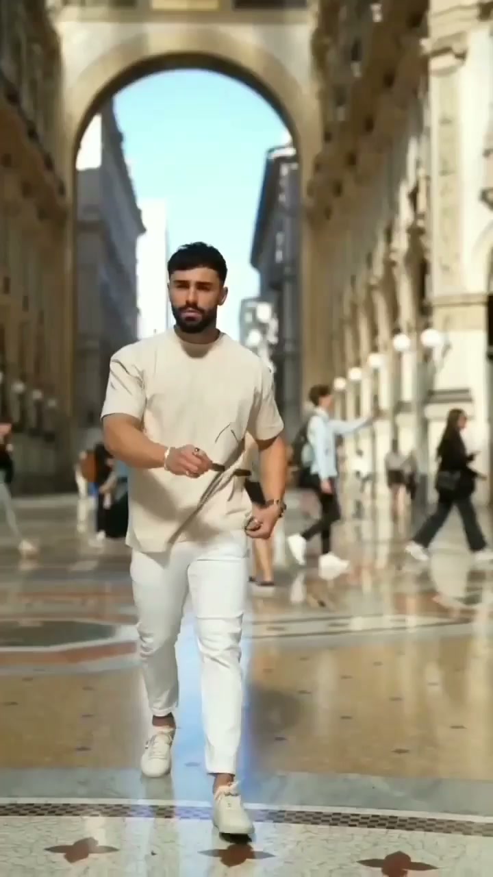 Man in a light beige T-shirt, fitted white trousers and white casual shoes in a covered city arcade