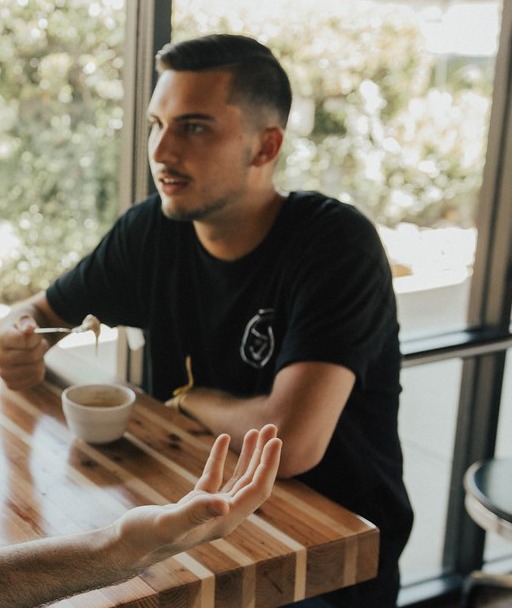A man in a black T-shirt listening at a wooden table with a speaker's gesturing hand in the foreground