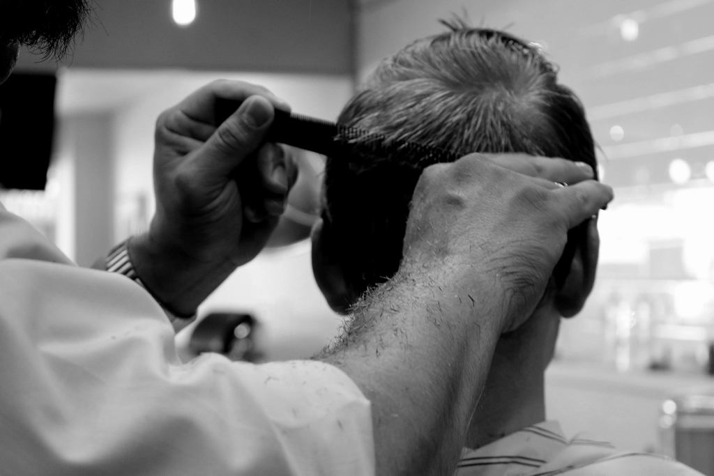 A man seated in a barber chair having his beard shaped with scissors and comb